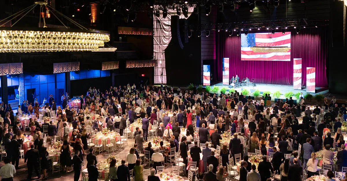 Wide room view of a Washington DC gala with guests, dinner tables, stage lighting, and an American flag backdrop